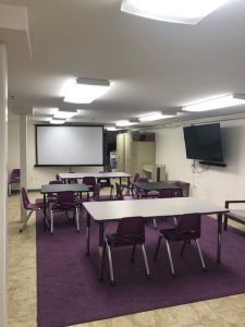 the program room features multiple tables and chairs, a large screen television, and a projection screen